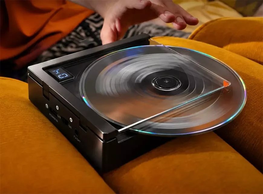 Lecteur CD portable Fosi Audio DISC Nano en finition noire posé sur un canapé jaune moutarde, CD en rotation sous un capot transparent, afficheur LED, entrée POWER IN, interrupteur et prise jack combinée Ω/OPT