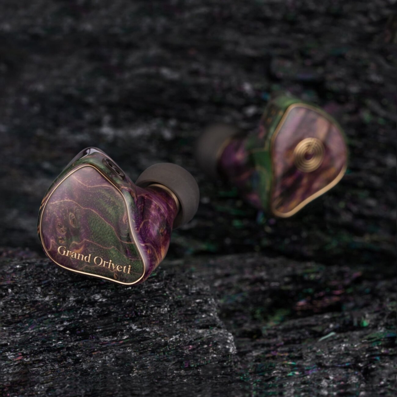 Intra-auriculaires Grand Oriveti Eminency posés sur une pierre sombre, façade en bois stabilisé violet et vert avec inscription Grand Oriveti dorée
