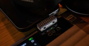Émetteur Bluetooth iFi UP Play Max branché en USB-C, avec ses boutons d'appairage et de codec, à côté d'un casque, compatible aptX Lossless et LDAC