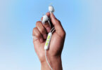 Écouteurs filaires Bose Noise Cancelling Wired Earbuds coloris blanc tenus en main, ailettes de stabilité, télécommande trois boutons vert anis et câble USB-C sur fond bleu ciel
