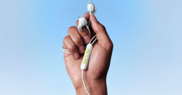 Écouteurs filaires Bose Noise Cancelling Wired Earbuds coloris blanc tenus en main, ailettes de stabilité, télécommande trois boutons vert anis et câble USB-C sur fond bleu ciel