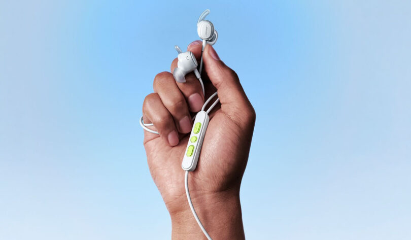 Écouteurs filaires Bose Noise Cancelling Wired Earbuds coloris blanc tenus en main, ailettes de stabilité, télécommande trois boutons vert anis et câble USB-C sur fond bleu ciel