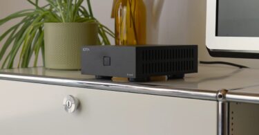 Ampli de puissance stéréo IOTAVX B:ound noir posé sur un meuble blanc, boîtier compact de 16,4 cm à ouïes d'aération latérales et bouton de mise en marche lumineux