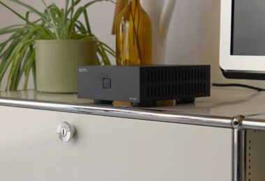Ampli de puissance stéréo IOTAVX B:ound noir posé sur un meuble blanc, boîtier compact de 16,4 cm à ouïes d'aération latérales et bouton de mise en marche lumineux