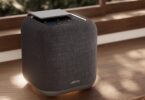 Enceinte connectée Jamo HYG 5 coloris gris foncé, cube habillé de tissu avec dessus en aluminium et commandes tactiles, posée sur une étagère en bois devant une fenêtre