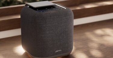 Enceinte connectée Jamo HYG 5 coloris gris foncé, cube habillé de tissu avec dessus en aluminium et commandes tactiles, posée sur une étagère en bois devant une fenêtre