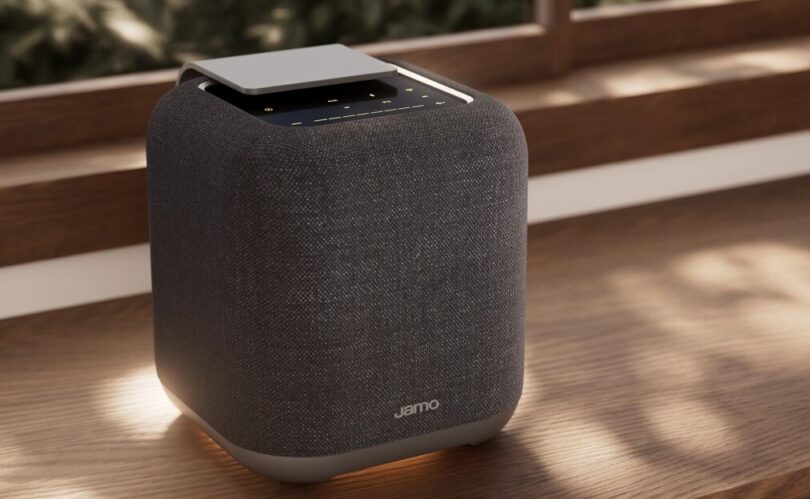 Enceinte connectée Jamo HYG 5 coloris gris foncé, cube habillé de tissu avec dessus en aluminium et commandes tactiles, posée sur une étagère en bois devant une fenêtre