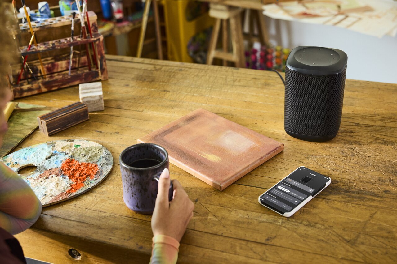 Enceinte compacte JBL Cove M1 noire posée sur une table en bois à côté d'un smartphone, d'une tasse et d'un carnet