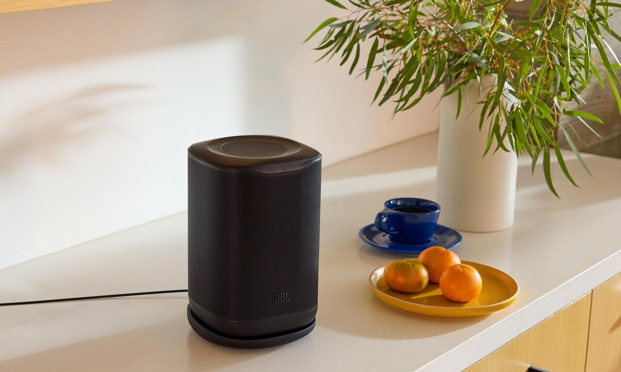 Enceinte portable JBL Cove P1 noire posée sur un meuble clair près d'une plante, d'une coupe de fruits et d'une tasse bleue