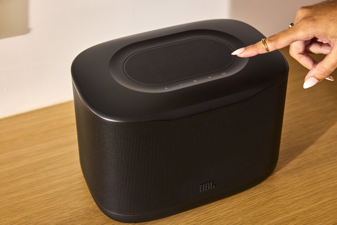 Main appuyant sur les commandes tactiles du dessus d'une enceinte JBL Cove X1 noire posée sur un meuble en bois