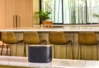 Enceinte multiroom Wi-Fi JBL Cove X1 noire posée sur un plan de travail en marbre dans une cuisine lumineuse avec tabourets de bar