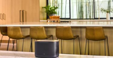 Enceinte multiroom Wi-Fi JBL Cove X1 noire posée sur un plan de travail en marbre dans une cuisine lumineuse avec tabourets de bar