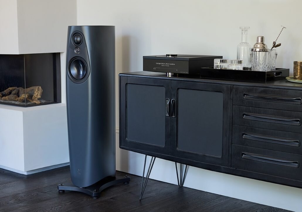 Enceinte Linn 343 finition Triton gris bleu posée sur son pied dans un salon à côté d'un meuble noir et d'un lecteur réseau Linn