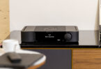 Amplificateur intégré MOON by Simaudio 341 noir posé sur un meuble hi-fi en bois, écran de 6,75 pouces affichant l'entrée en français et gros bouton de volume