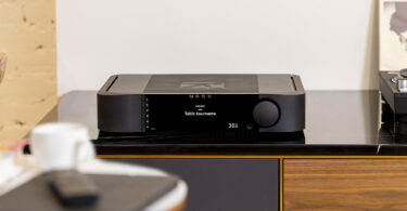 Amplificateur intégré MOON by Simaudio 341 noir posé sur un meuble hi-fi en bois, écran de 6,75 pouces affichant l'entrée en français et gros bouton de volume