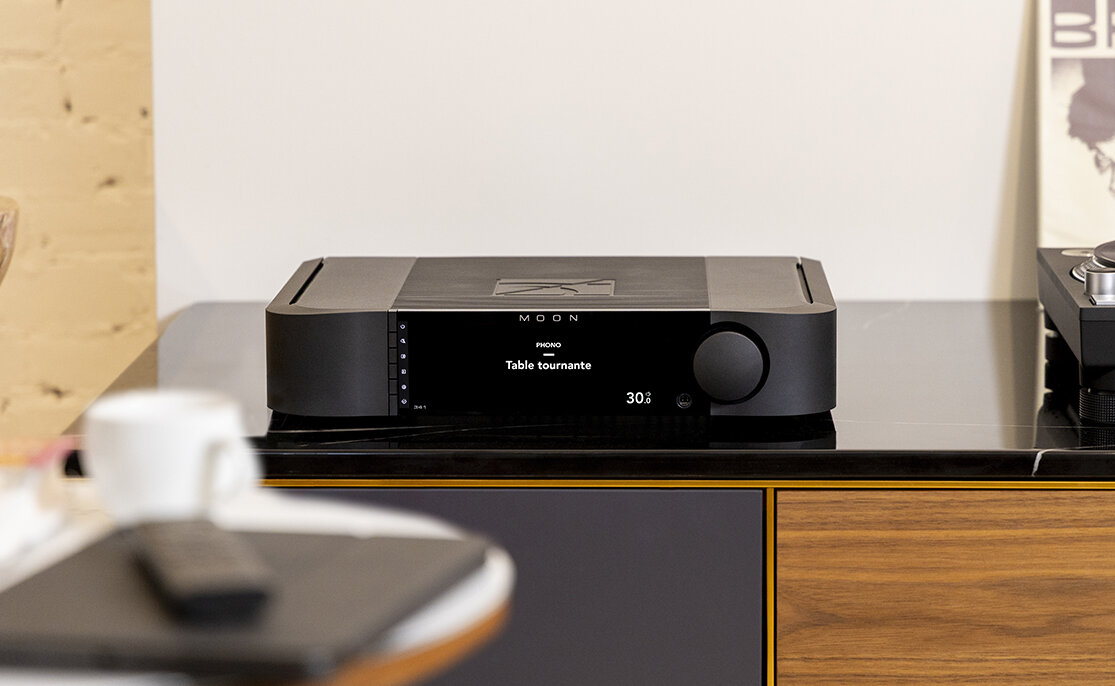 Amplificateur intégré MOON by Simaudio 341 noir posé sur un meuble hi-fi en bois, écran de 6,75 pouces affichant l'entrée en français et gros bouton de volume