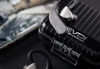 Crochets d'oreille Bluetooth Moondrop EVO 2 (EVO II) gris anthracite reliés à des écouteurs intra-auriculaires argentés, posés sur l'étui de charge renforcé noir à rainures marqué EVO II, smartphone en arrière-plan