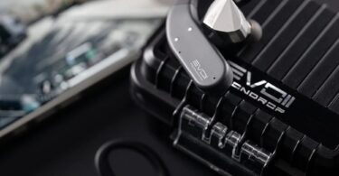 Crochets d'oreille Bluetooth Moondrop EVO 2 (EVO II) gris anthracite reliés à des écouteurs intra-auriculaires argentés, posés sur l'étui de charge renforcé noir à rainures marqué EVO II, smartphone en arrière-plan
