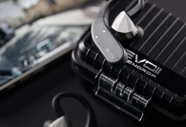 Crochets d'oreille Bluetooth Moondrop EVO 2 (EVO II) gris anthracite reliés à des écouteurs intra-auriculaires argentés, posés sur l'étui de charge renforcé noir à rainures marqué EVO II, smartphone en arrière-plan
