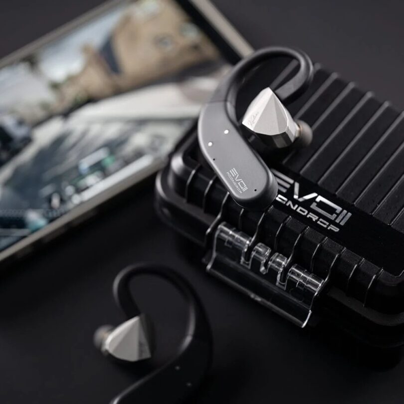Crochets d'oreille Bluetooth Moondrop EVO 2 (EVO II) gris anthracite reliés à des écouteurs intra-auriculaires argentés, posés sur l'étui de charge renforcé noir à rainures marqué EVO II, smartphone en arrière-plan