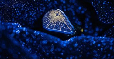 Écouteur intra-auriculaire Moondrop Starfield 3 posé sur un tissu bleu scintillant, plaque de façade bleu nuit pailletée avec étoile et rayons dorés