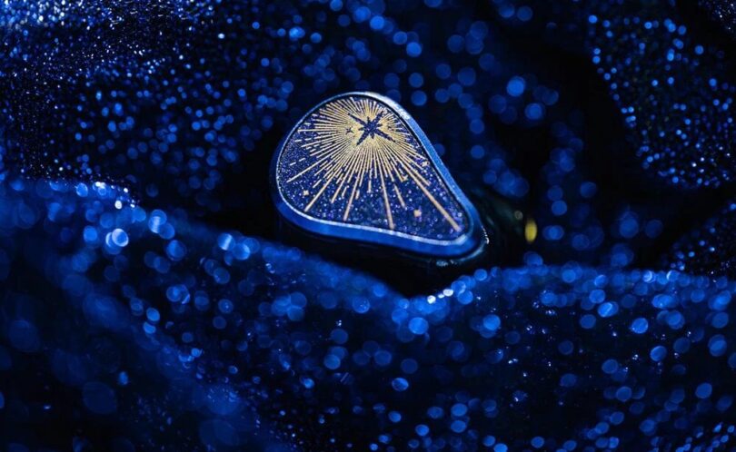 Écouteur intra-auriculaire Moondrop Starfield 3 posé sur un tissu bleu scintillant, plaque de façade bleu nuit pailletée avec étoile et rayons dorés