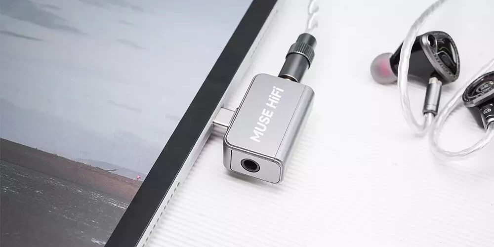 Dongle DAC Muse HiFi U3 en aluminium argent branché sur le port USB-C d'une tablette, avec des écouteurs intra-auriculaires reliés à sa prise jack 3,5 mm