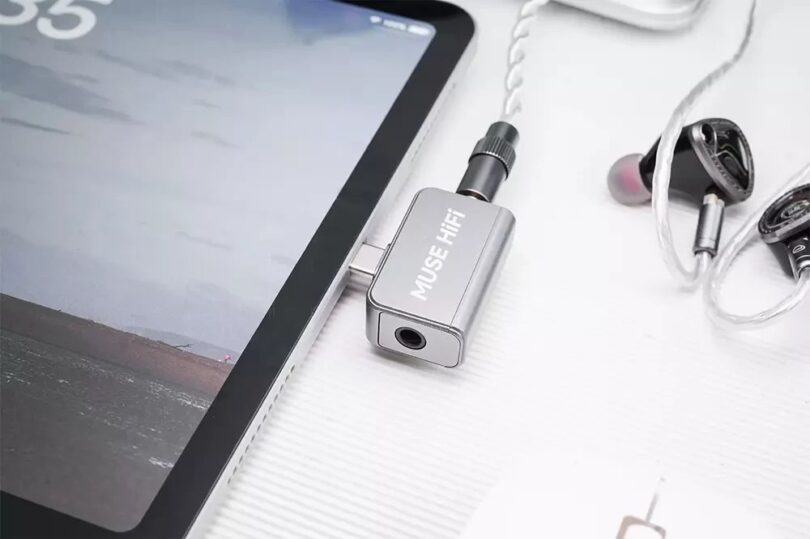 Dongle DAC Muse HiFi U3 en aluminium argent branché sur le port USB-C d'une tablette, avec des écouteurs intra-auriculaires reliés à sa prise jack 3,5 mm