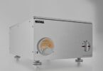 Amplificateur de puissance stéréo Nagra AMP II-S en aluminium brossé argent vu de trois quarts, avec Modulomètre à aiguille rétroéclairé et sélecteur rotatif en façade, posé sur pieds découplés