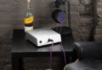 Ampli casque Orchard Audio Valencia en finition argent posé sur un meuble noir, câble de casque violet branché en façade, casque sur son support devant un mur de briques