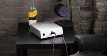 Ampli casque Orchard Audio Valencia en finition argent posé sur un meuble noir, câble de casque violet branché en façade, casque sur son support devant un mur de briques