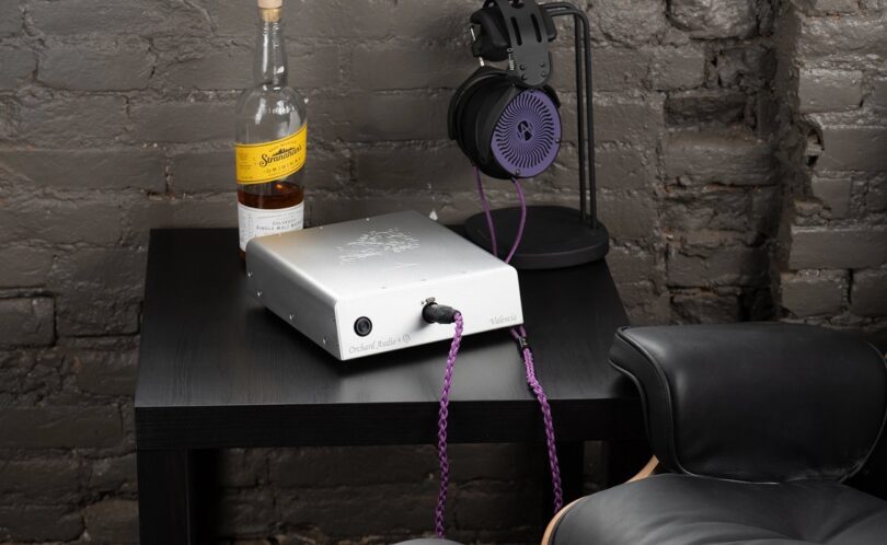 Ampli casque Orchard Audio Valencia en finition argent posé sur un meuble noir, câble de casque violet branché en façade, casque sur son support devant un mur de briques