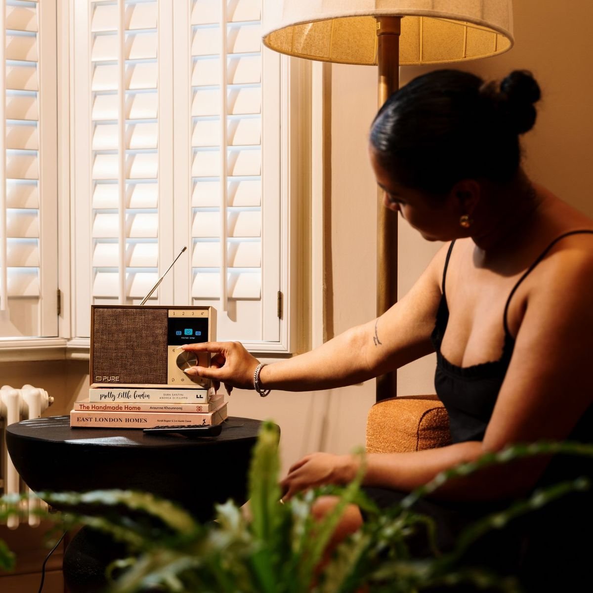 Une personne règle la molette d'une radio Pure Evoke One en bois posée sur une pile de livres dans un salon, antenne télescopique déployée, lampe et plantes autour