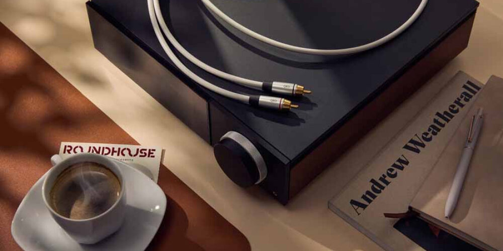 Câble de modulation RCA QED Performance Audio à gaine TPE gris clair et fiches gris argent marquées QED à pointes plaquées or, posé sur un appareil hi-fi noir à bouton de volume moleté