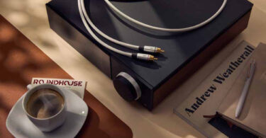 Câble de modulation RCA QED Performance Audio à gaine TPE gris clair et fiches gris argent marquées QED à pointes plaquées or, posé sur un appareil hi-fi noir à bouton de volume moleté