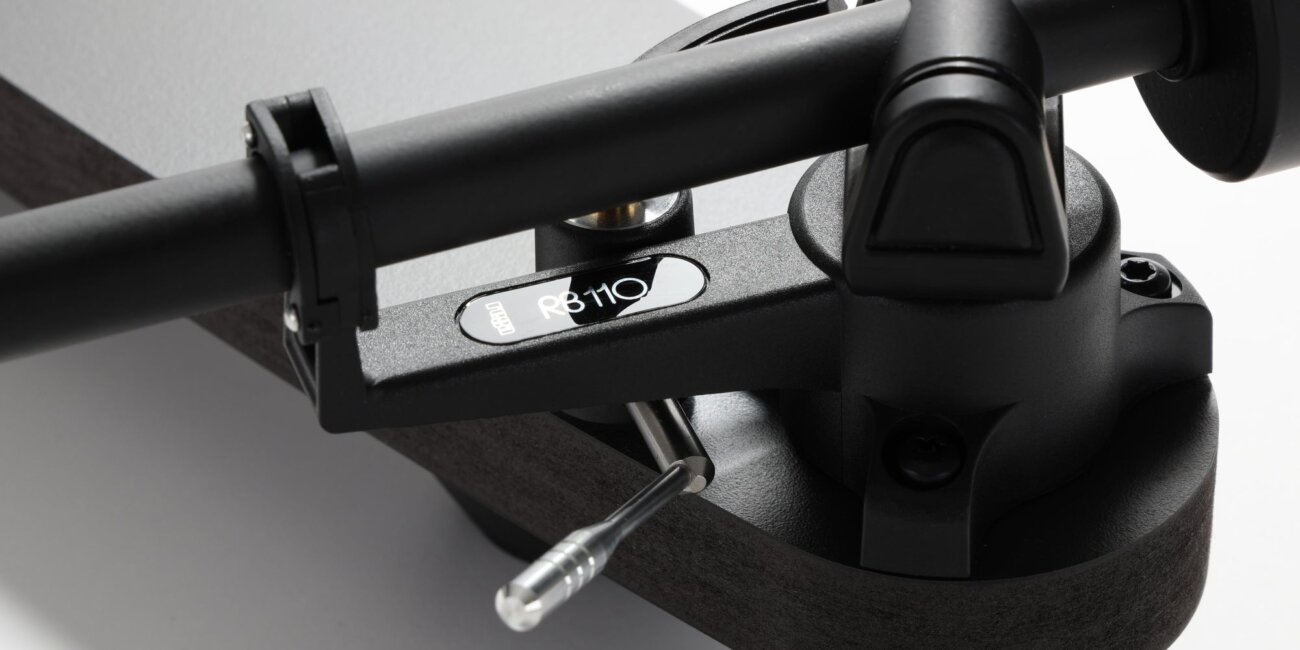 Gros plan sur le bras Rega RB110 de la platine Replay, avec son support, son lève-bras et sa plinthe noire