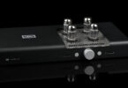 réamplificateur symétrique Schiit Freya 2 en finition noire, vu de trois quarts, avec ses quatre lampes octales émergeant de la grille ajourée du capot, son bouton de volume central argenté et ses deux afficheurs en façade