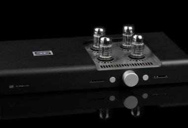 réamplificateur symétrique Schiit Freya 2 en finition noire, vu de trois quarts, avec ses quatre lampes octales émergeant de la grille ajourée du capot, son bouton de volume central argenté et ses deux afficheurs en façade