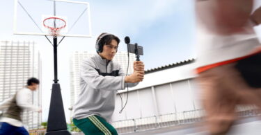 Vidéaste filmant un match de basket au smartphone équipé du micro spatial Sony ECM-AX10 à sphère noire, avec écoute de contrôle au casque branché sur la sortie mini-jack du micro