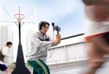 Vidéaste filmant un match de basket au smartphone équipé du micro spatial Sony ECM-AX10 à sphère noire, avec écoute de contrôle au casque branché sur la sortie mini-jack du micro