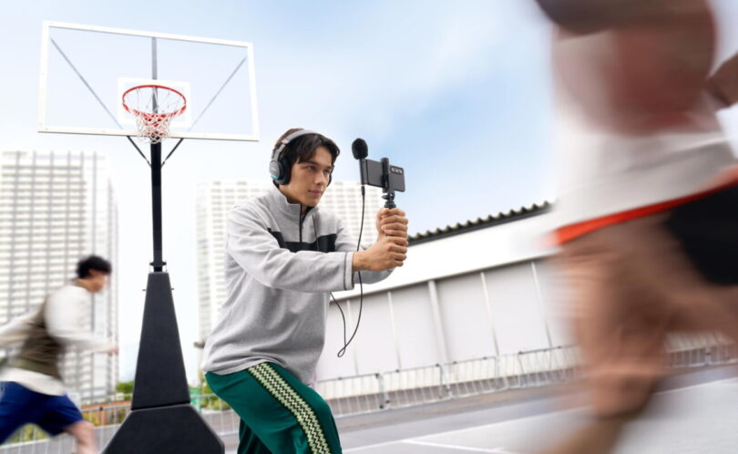 Vidéaste filmant un match de basket au smartphone équipé du micro spatial Sony ECM-AX10 à sphère noire, avec écoute de contrôle au casque branché sur la sortie mini-jack du micro